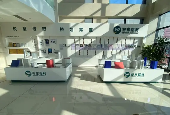 Yoverd Aluminum exhibition hall product display showcasing various aluminum profiles and alloy extrusions (silver profiles, red cylinder, blue panels, etc.), with company honor wall, About Us introduction, and timeline in the background, and YOVERD ALUMINIUM brand logo at the bottom